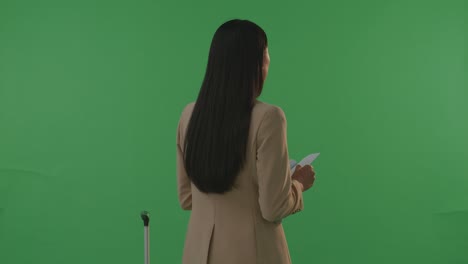vista trasera mujer de negocios asiática viajera con equipaje mirando el pasaporte y mirando a su alrededor mientras está de pie en el estudio de fondo de pantalla verde, durante la hora de la puesta del sol