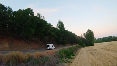 Camper-van-travels-down-a-paved-road-near-a-field-and-trees-at-sunset