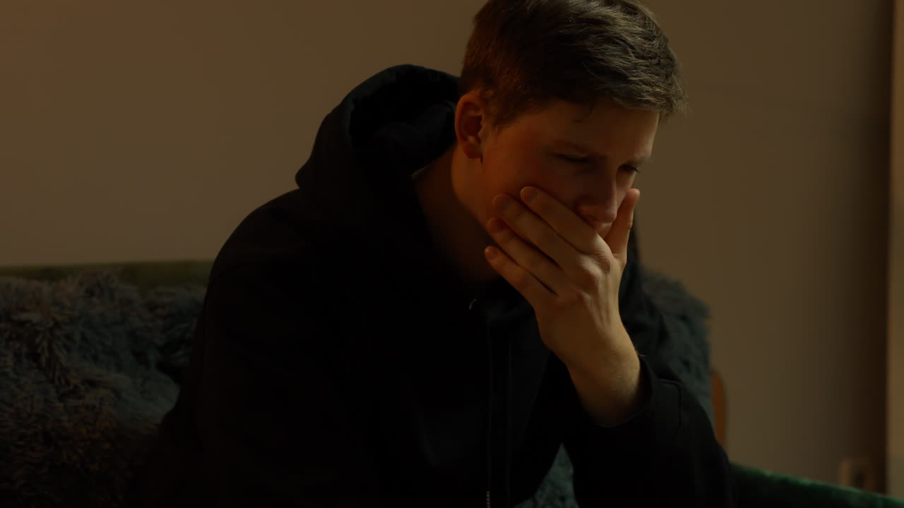 Jovem cobrindo a boca com a mão, expressando sentimentos de tristeza, choque e pesar, enfrentando dor emocional, preocupação e desespero em um momento de silêncio e profunda reflexão, em um quarto com pouca luz