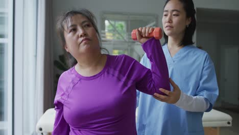 Asian-female-physiotherapist-helping-female-patient-exercise-arms-with-dumbbells-at-surgery