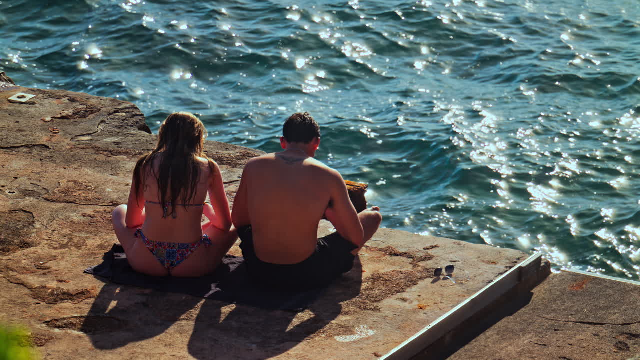 Villefranche sur Mer, France - March 28, 2025: A man and a woman sitting and sunbathing on a stone pier near the sea