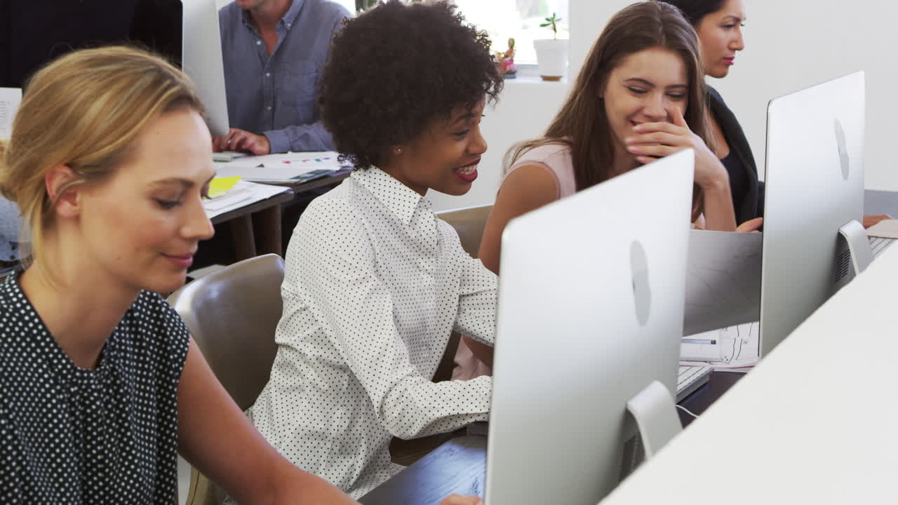 Two happy women discuss work at computer in open plan office