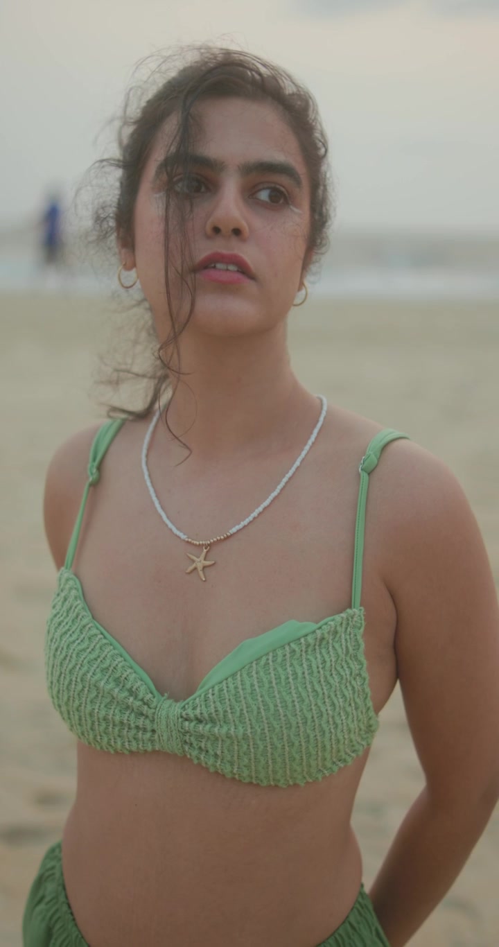 Una giovane donna in bikini verde sta in piedi sferzata dal vento su una spiaggia, con lo sguardo perso all'orizzonte.
