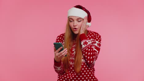 Frau-Im-Stilvollen-Weihnachtspullover-Schaut-Auf-Das-Smartphone-Display-Und-Freut-Sich-Aufrichtig-über-Den-Sieg,-Den-Erfolg-Und-Das-Glück