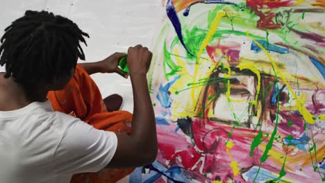 Overhead-view-of-african-american-male-artist-pouring-paint-with-a-paint-brush-on-canvas