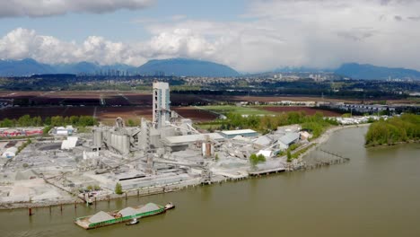 Sand-Barge-Sailing-In-The-Fraser-River-With-Cement-Factory-In-Lulu-Island,-Richmond,-BC,-Canada