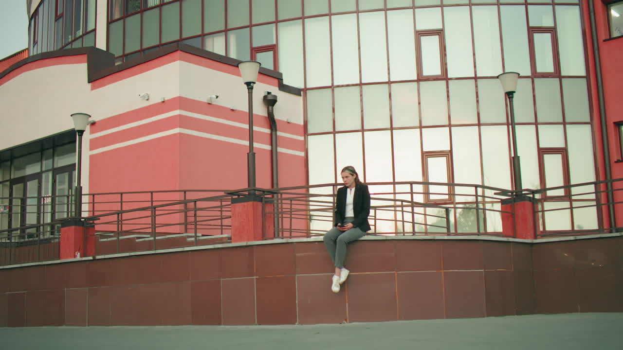 Calm lady in black blazer seated on fence with legs crossed at ankle holding phone, looking down in peaceful urban setting with car reflection on sleek tiles and modern glass building background