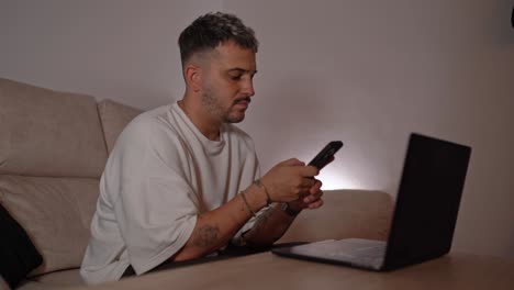 side view of man sitting on sofa typing on mobile phone with laptop open on table in home setting, under soft ambient lighting