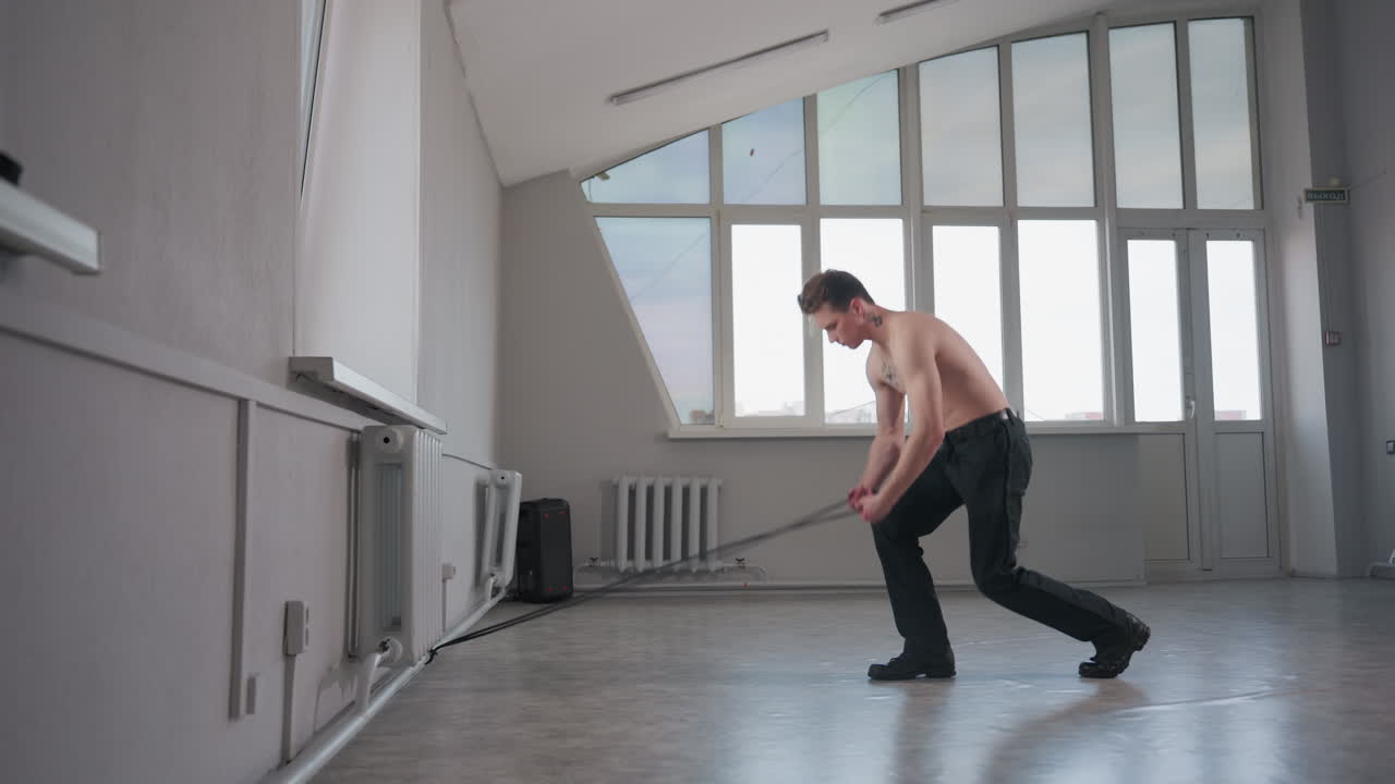 shirtless man with neck tattoo walks across bright studio floor to thick rope lying on ground, bends to tie rope around wrist and begins dragging it back