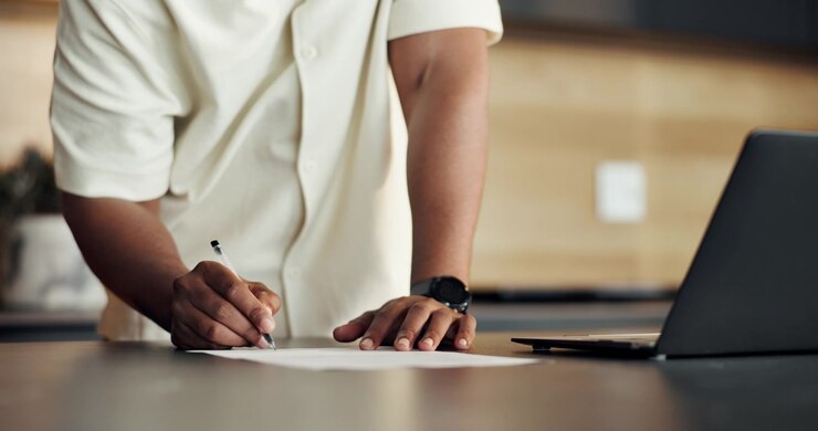Person Signing Document on Desk Next to Laptop
