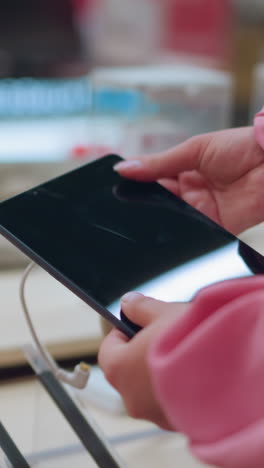 dama vista desde atrás recogiendo una tableta de su soporte en una tienda, la tableta está conectada con un cable de seguridad, mientras que otras tabletas están dispuestas cuidadosamente en la mesa, ella inspecciona el dispositivo de cerca
