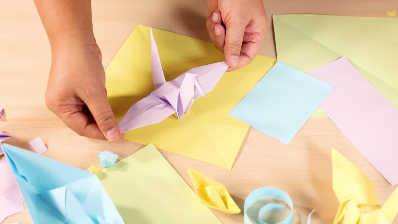 Person folds origami crane with pastel paper, overhead view, bright lighting, steady camera