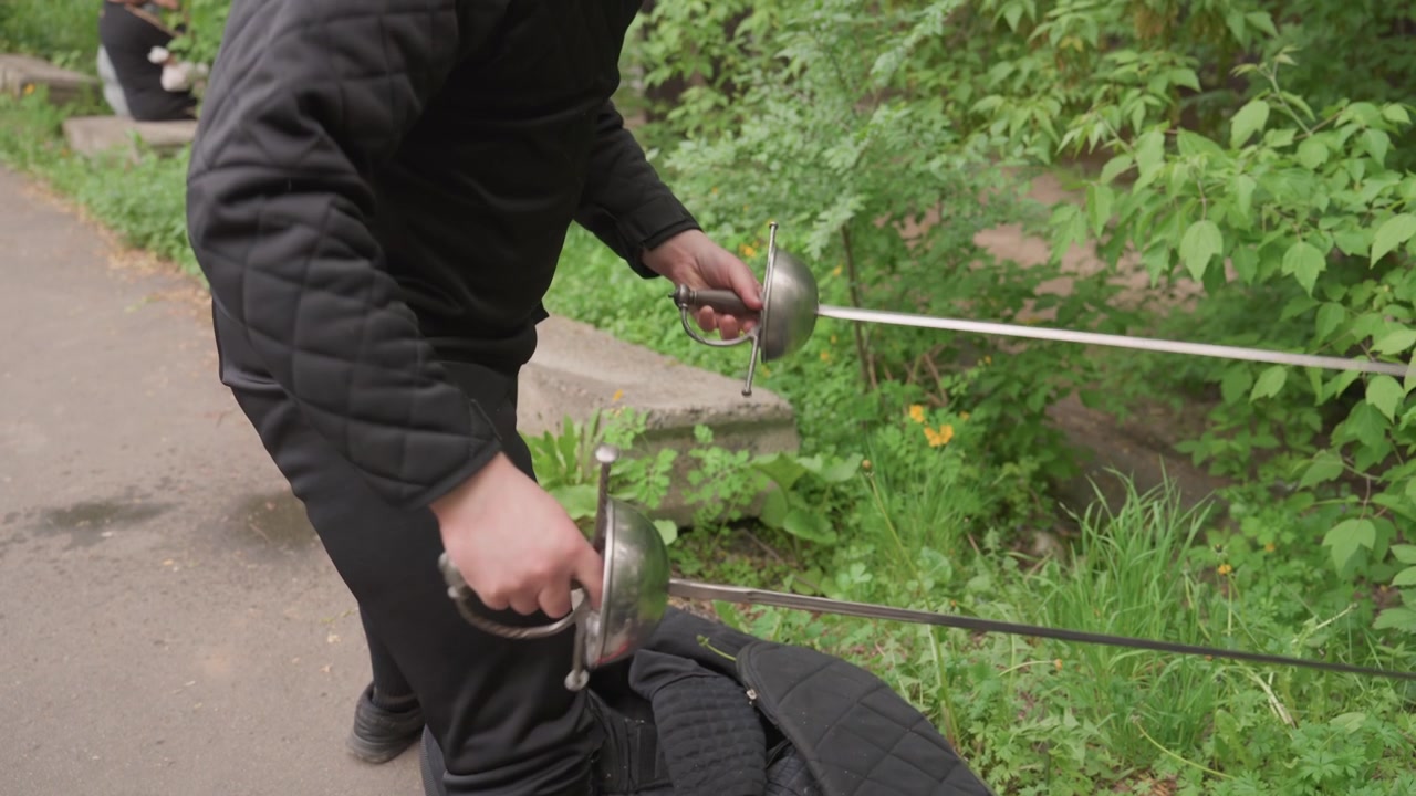 Close up of man in black fencing gear handling two swords on pavement with greenery background. man adjusts and moves metallic swords demonstrating their grip from various angles