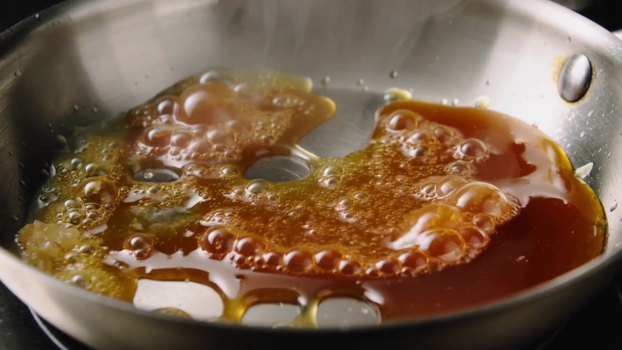 Heating dark amber syrup, bubbling and spreading on stainless-steel skillet with rivets, reducing