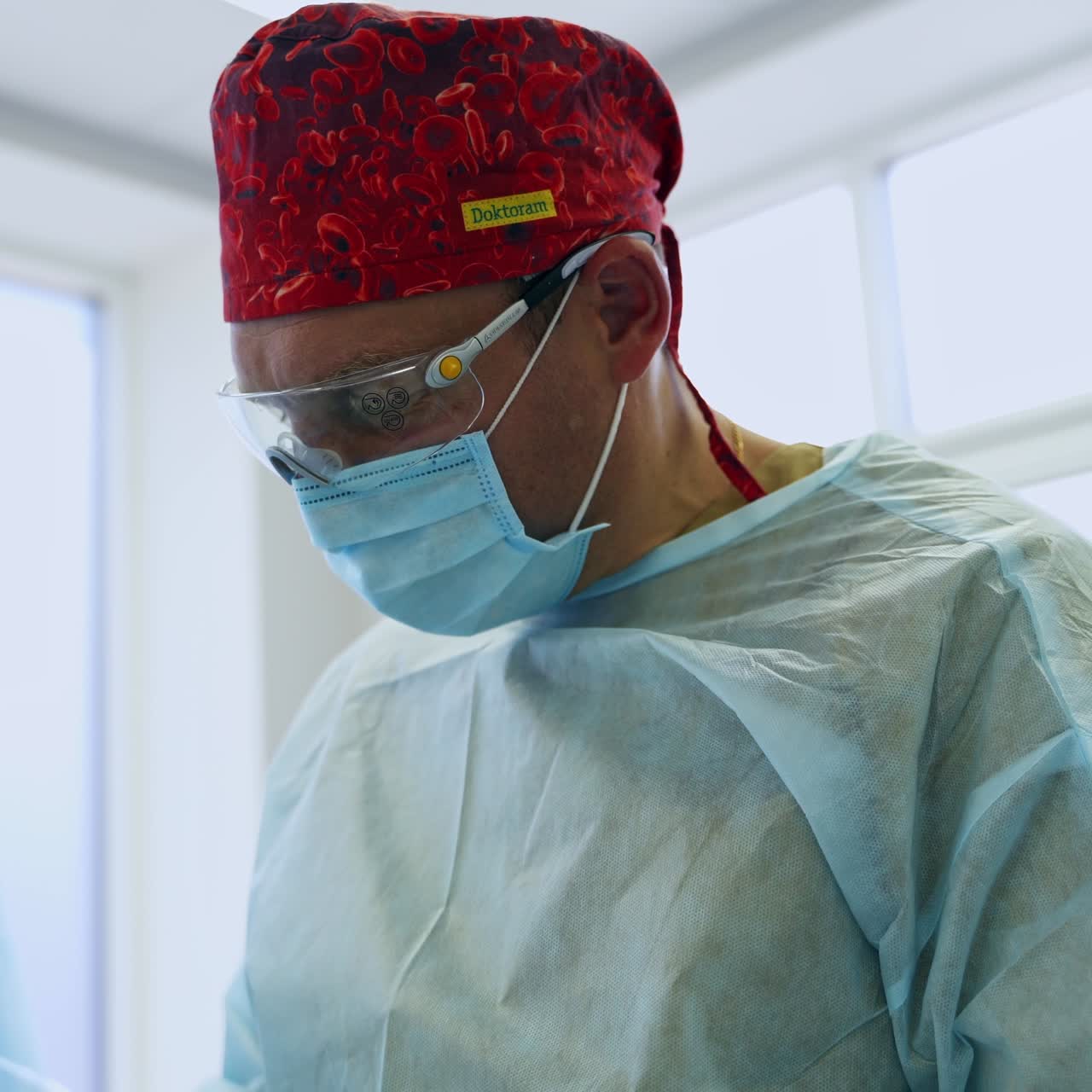 Adult male surgeon wearing uniform, red cap, mask and glasses. Medical professional being busy with operation. Low angle view