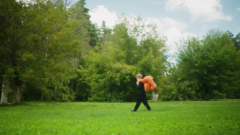 Side view of young boy holding large teddy bear behind his back while walking through open green park under clear sky, surrounded by trees and enjoying quiet outdoor moment in nature