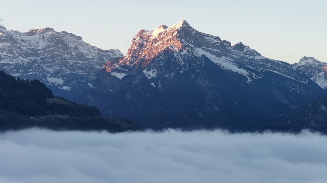 Vista aérea incrível do maciço Mürtschenstock, que se eleva sobre um denso mar de neblina. A luz dourada do sol atinge o pico nevado suíço, criando uma vista alpina divina sobre o lago Walensee