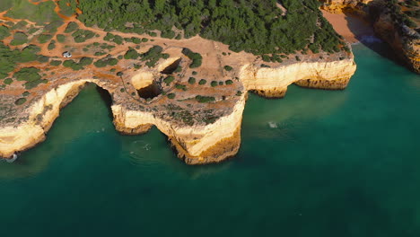 drone shot above algarve cliffs with blue and green water, trees inland, a hidden sandy beach, and a campercar parked on the land. stunning view of nature, coastline. .