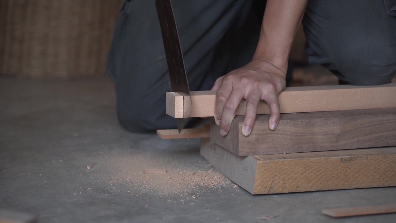 Craftsman cutting wooden board with hand saw on stacked timber, producing fine sawdust during woodworking process inside carpentry workshop