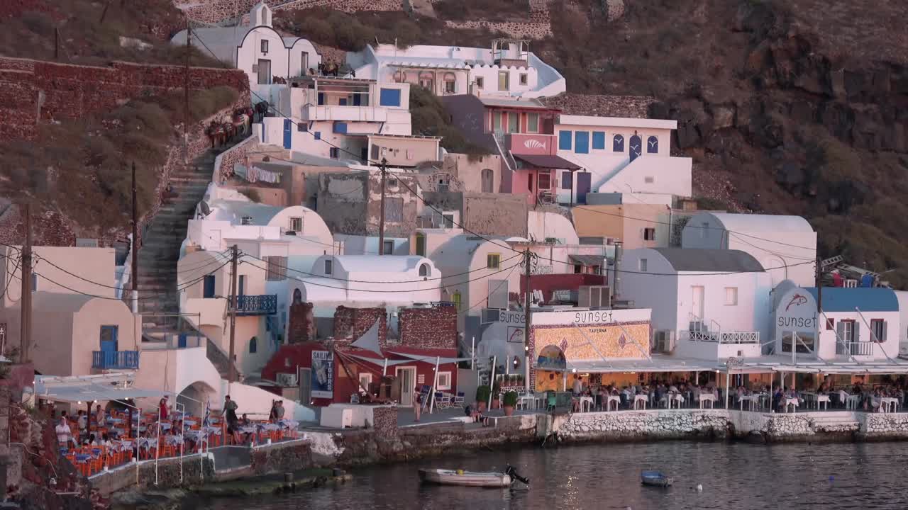 Vista panorâmica de uma vila costeira no penhasco com edifícios brancos, restaurantes à beira-mar e pequenos barcos ao longo da costa, sob a luz quente do entardecer em Santorini, Grécia.