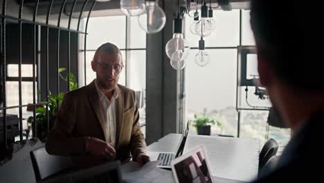 A-blond-man-in-glasses-and-a-brown-suit-proposes-his-idea-to-his-colleague-using-papers.-Employees-communicating-in-a-modern-office.-Two-men-sitting-at-a-table-and-talking