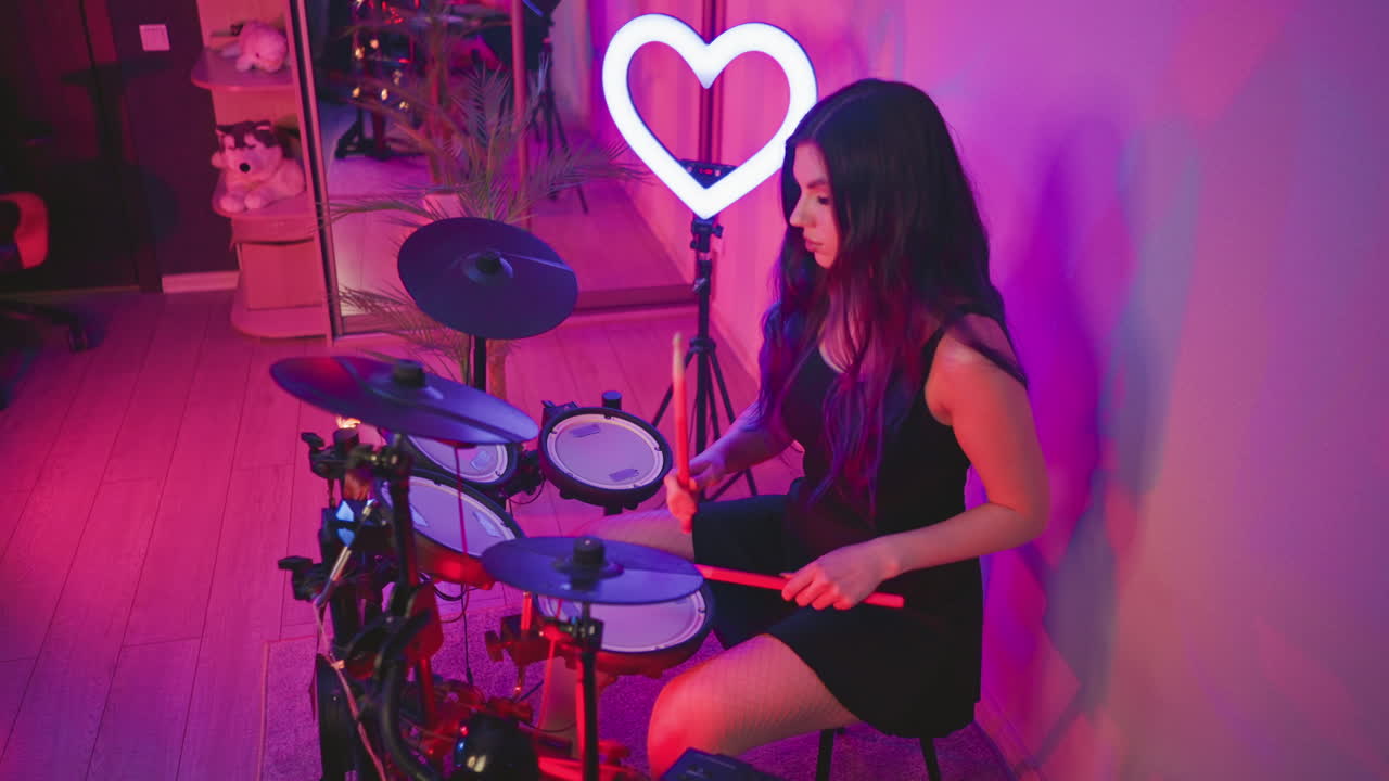 Drummer girl practicing on electronic drum kit under purple lighting in home studio, with glowing heart-shaped light and mirror reflecting room details in background