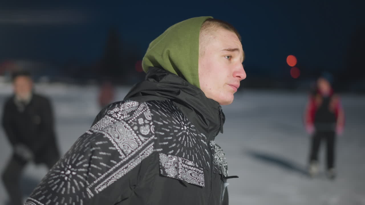 young man gliding on outdoor ice rink during cold night wearing patterned black jacket and green hoodie with serious facial expression bright lights and blurred snowy background