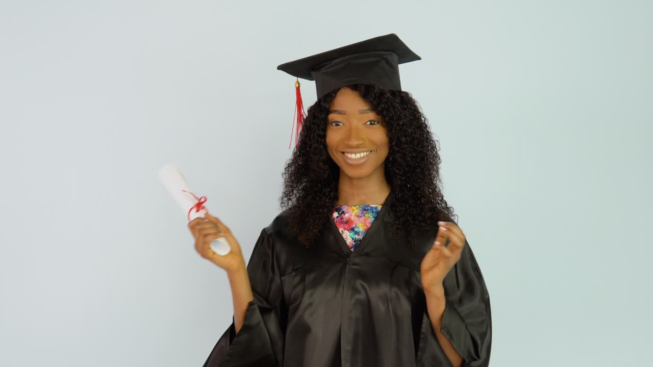 una joven afroamericana con una bata negra y un sombrero de maestro se pone erguida y agita el diploma de graduación en su mano. mira a la cámara. retrato de media altura