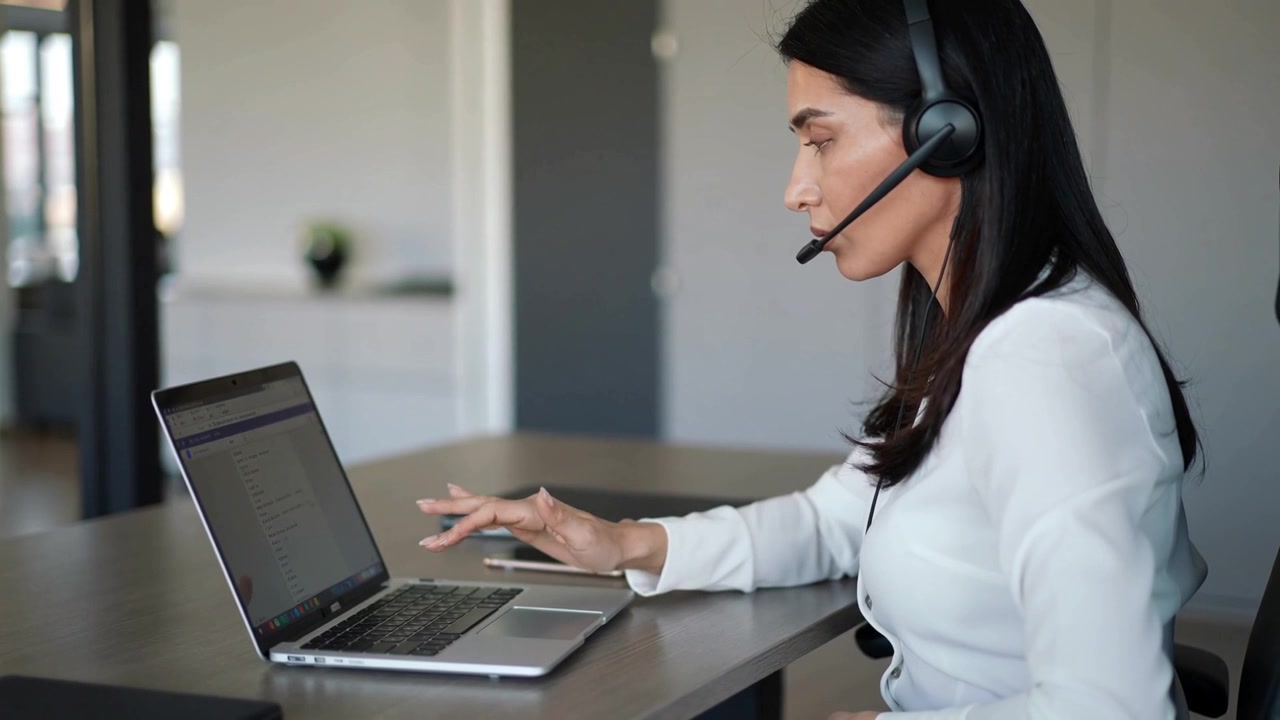 Ricevendo una chiamata, una donna con maglietta bianca e cuffie si sposta e digita mentre spiega al laptop a casa, con spazio per il testo.