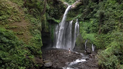 Majestic-aerial-shot-of-girl-raising-hands-up-in-the-air-with-flowing-Sendang-Gile-waterfall-in-background