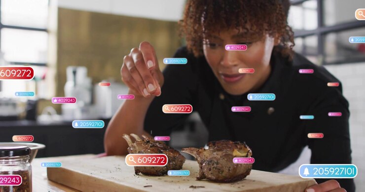 Chef picking up metal shaker and evenly seasoning lamb racks while digital food tags rising