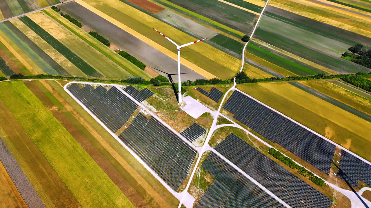 Wind turbine with red stripes rotates in the stripy field. Solar panel site located around the wind mills. Aerial view