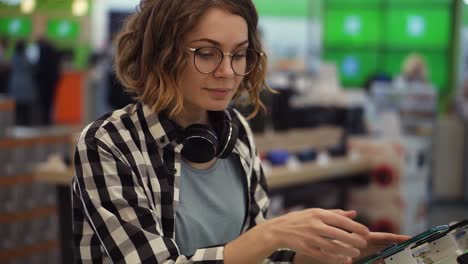 curly woman with headphones on neck standing at the counter with mobile phones in casual clothes choosing a new smartphone in a modern electronics store