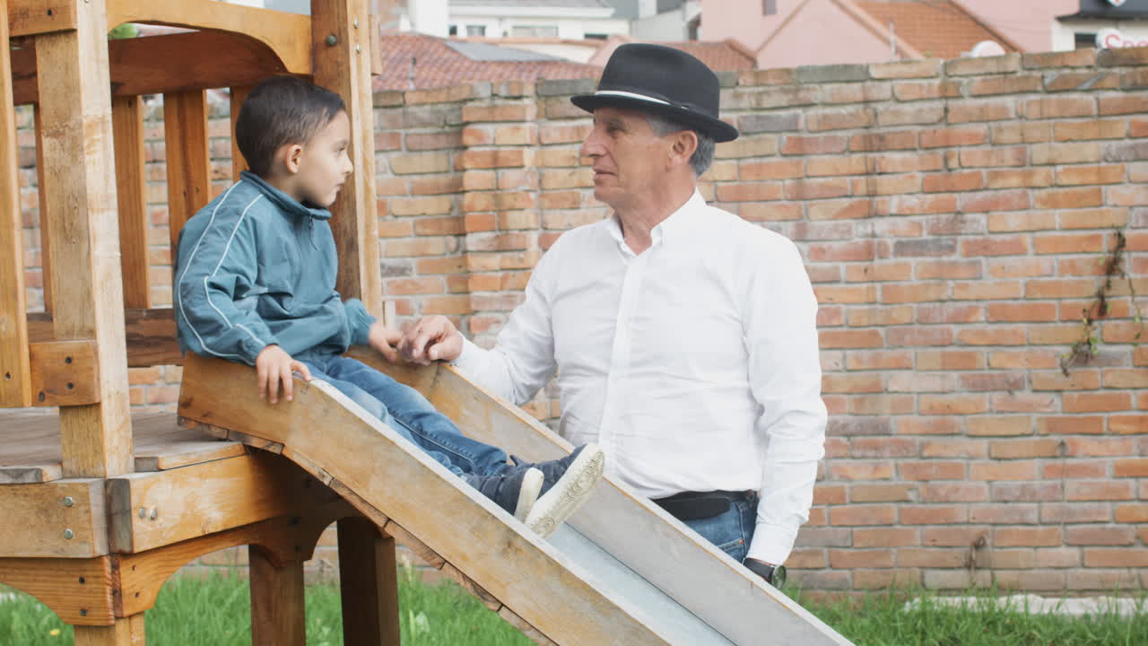 A tender moment between grandfather and grandson as they play together in a wooden playhouse with a slide in the backyard.