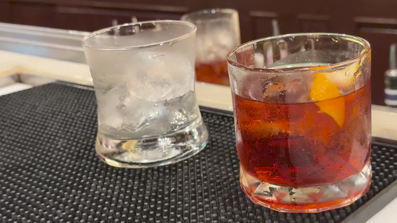 Bartender slides cocktail glass with ice beside Negroni on bar mat, warm indoor lighting, close-up