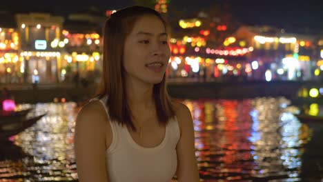 medium shot of asian girl at hoi an ancient town riverfront at night - blurry boats on river and colorful lights in background