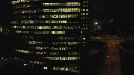 panning right aerial shot in the dark, evening of an office building with lights on working over hours, and traffic with cars