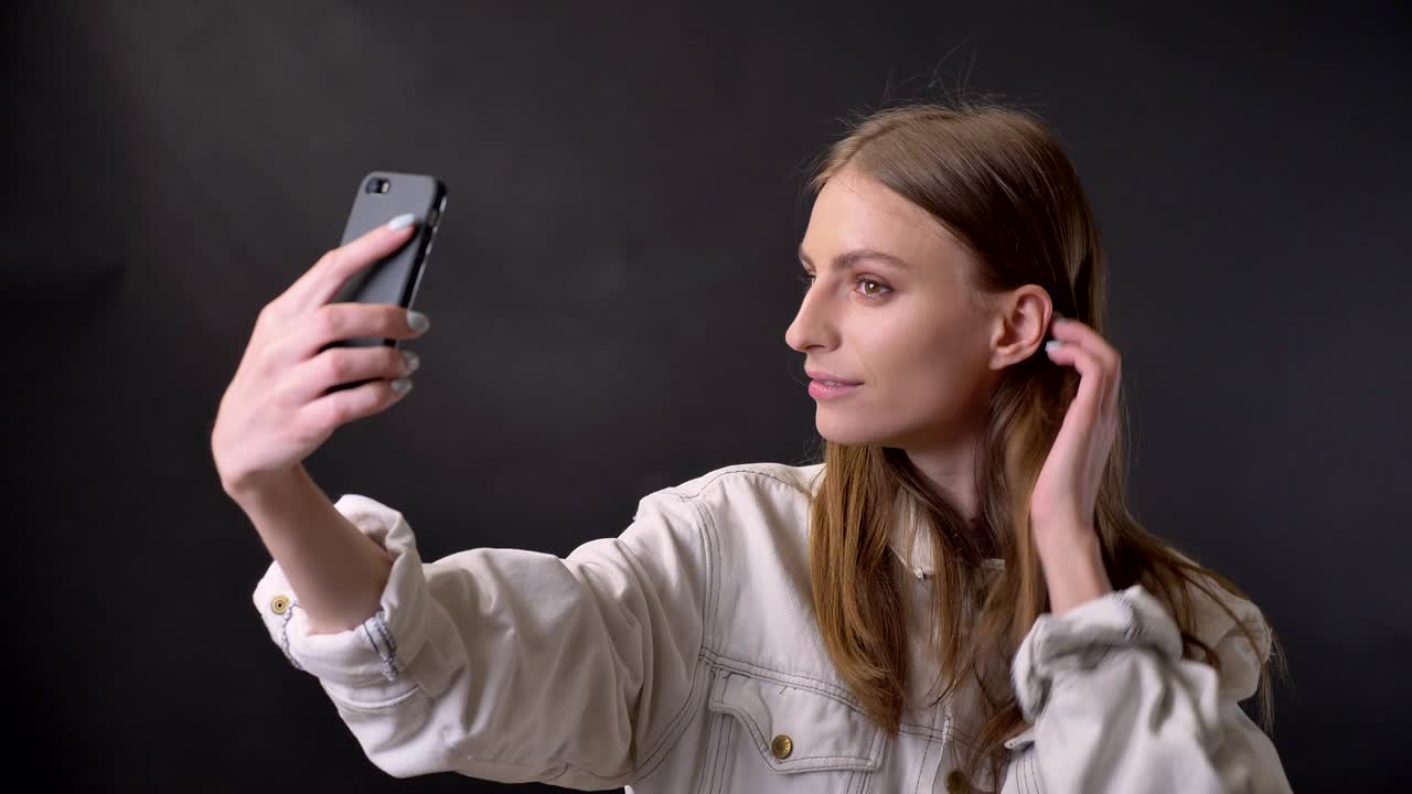 chica joven está haciendo selfie en el teléfono inteligente, tocando el cabello, concepto de comunicación, fondo gris
