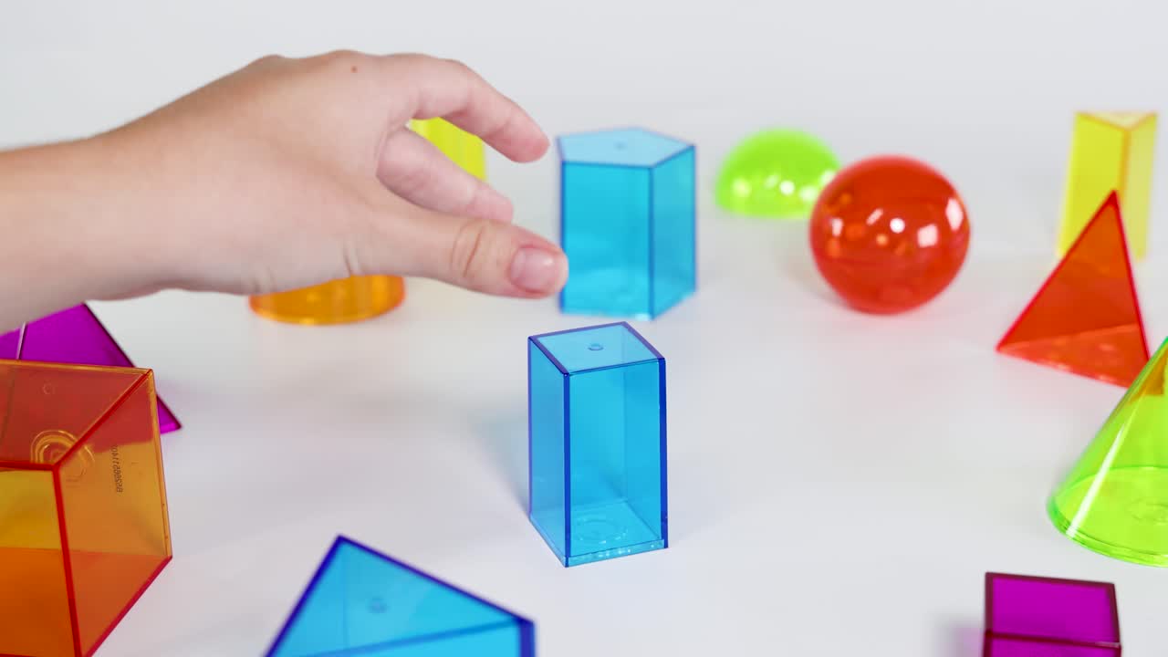 Hands interact with colorful 3D geometric shapes on a white surface, highlighting educational play and spatial awareness