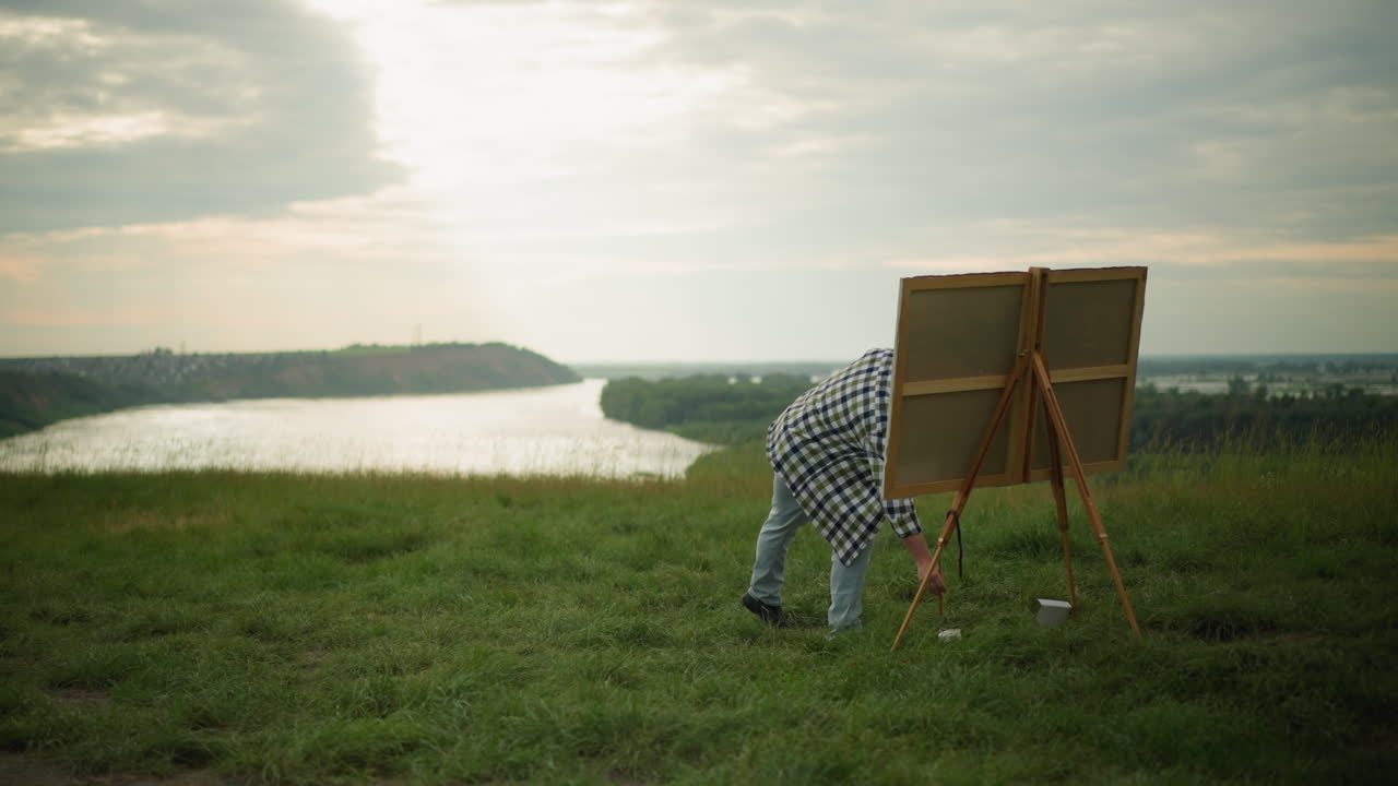 An artist wearing a black hat, checkered shirt, and jeans is painting on a large canvas in a peaceful grass field by a serene lake