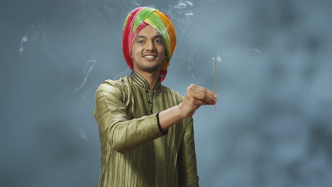Young-happy-indian-man-in-turban-and-traditional-clothes-smiling-at-camera-and-holding-incense-stick