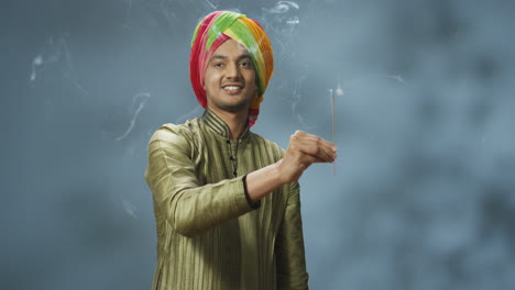 young happy indian man in turban and traditional clothes smiling at camera and holding incense stick