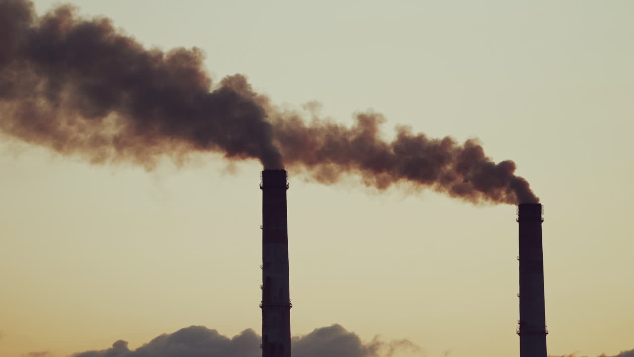 Large industrial pipes pollute the atmosphere with smoke. Two big pipes with thick smoke in the air on the grey evening sky background.