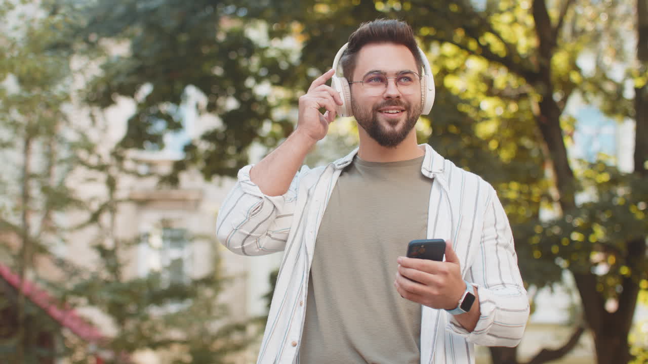 Happy young man putting on headphones listening favorite energetic music in smartphone on street