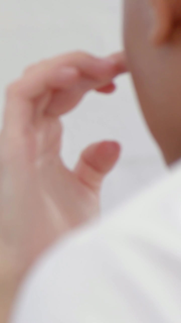 Vertical video: Sensing itch man raising right hand to ear in room, touching temple in white shirt