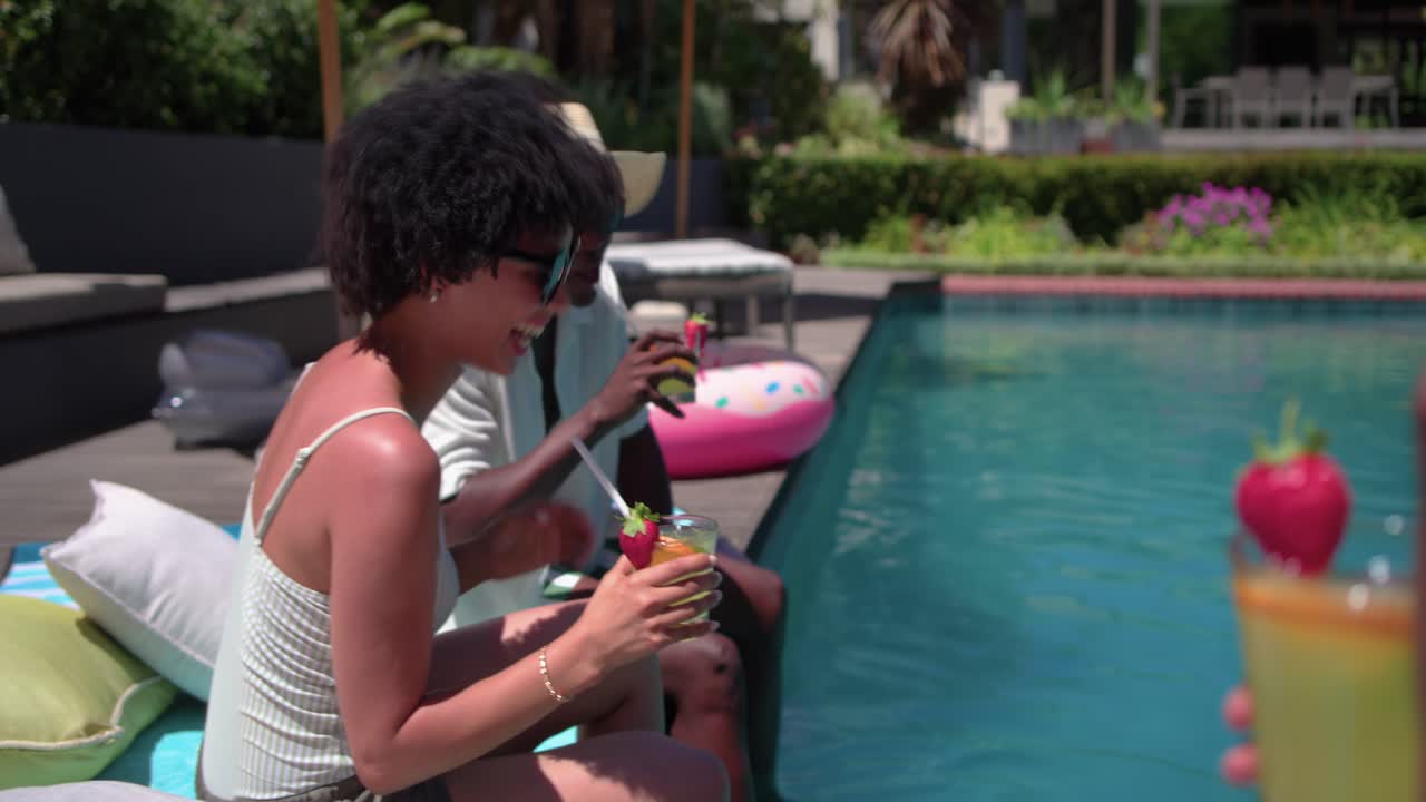 Friends sitting poolside standing woman coming offering drinks man in hat toasting and sipping