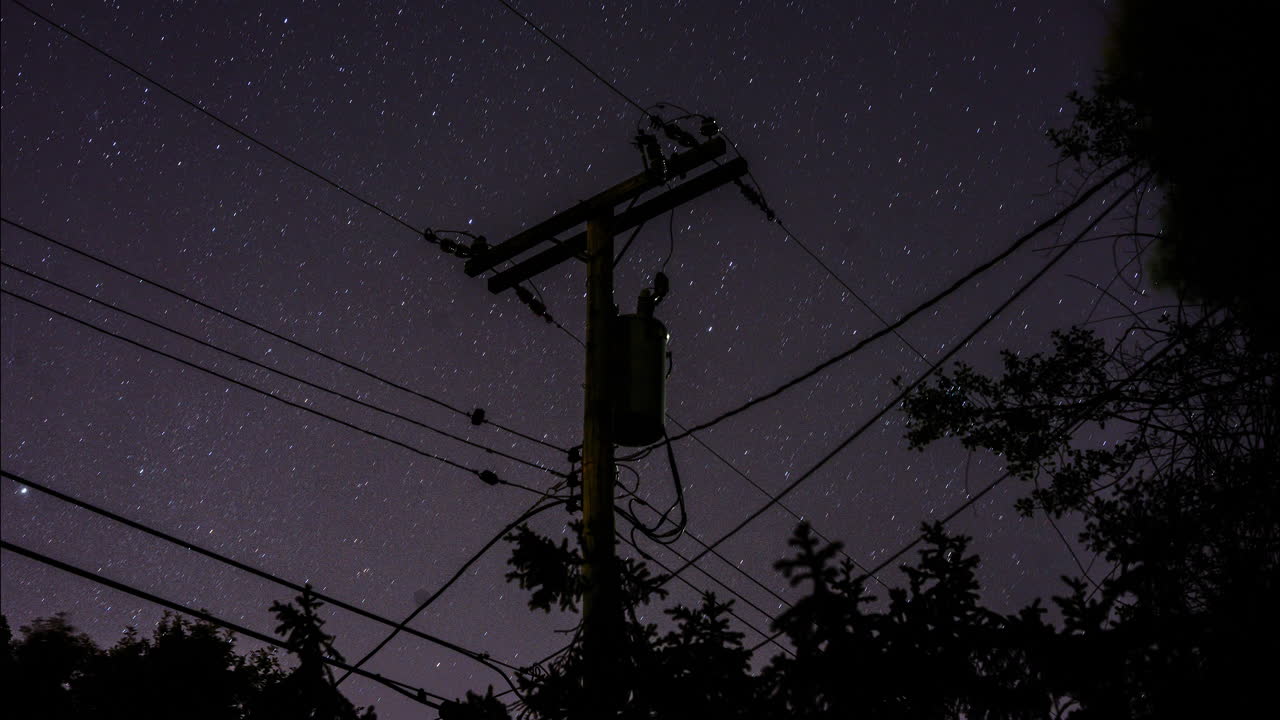 A time-lapse of the stars rotating in the sky behind a silhouetted suburban telephone pole and power lines and trees in the night sky