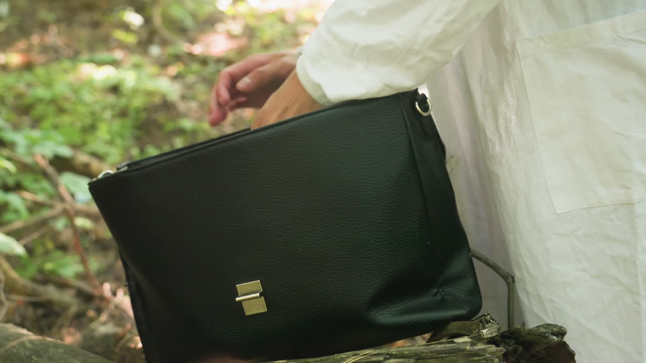 Close view of black backpack placed on old tree stump as hand carefully places jotter and pen inside during outdoor preparation in blurred forest environment surrounded by greenery and natural light