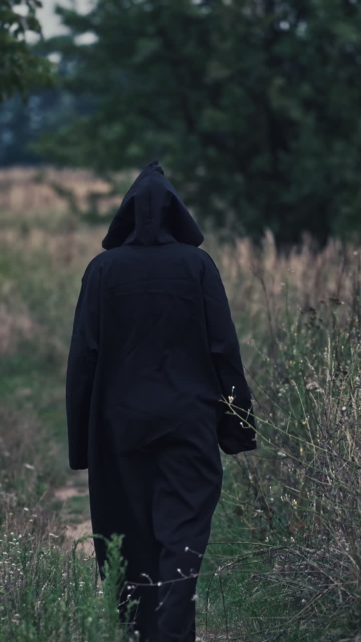 Backside view of witch on the field. Black death goes slowly away along the path on nature background. Vertical video