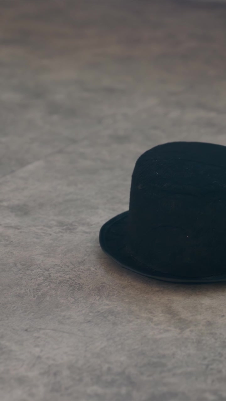 Primo piano di una bombetta nera appoggiata su una superficie grigia e texturizzata. Il cappello rimane fermo mentre la telecamera si sposta lentamente a sinistra, rivelando. Ambiente interno con dettagli di sfondo neutri, con la messa a fuoco.